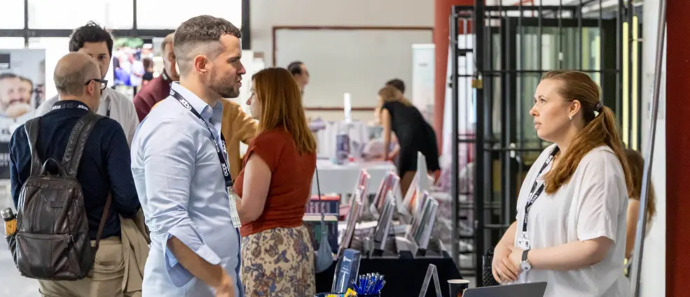 People networking in the exhibition area at ECPR conference. A man in a blue shirt talks to a woman in a white top, both wearing lanyards. Others converse in the background.